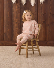 Load image into Gallery viewer, A toddler girl with blonde curly hair wearing a light pink long-sleeve pajama set with white polka dots sits on a small rustic wooden chair in front of a dark wood plank wall.
