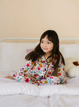 Load image into Gallery viewer, A young girl with dark hair sits smiling on a white bed wearing a pajama set with a colorful Halloween print of pumpkins, ghosts, and spiders. The background is a soft beige wall.
