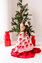 Load image into Gallery viewer, Seated child looking over their shoulder in a blush pink knit cardigan with red bows, layered over a red dress.
