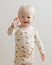 Load image into Gallery viewer, A smiling toddler with blonde hair wearing a cream-colored long-sleeve pajama set with a pumpkin and acorn pattern, waving at the camera against a plain light background.
