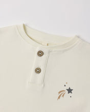 Load image into Gallery viewer, Close-up of a cream-colored children's henley shirt featuring a three-button placket and a small embroidered star and burst graphic on the chest.
