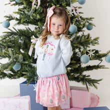 Load image into Gallery viewer, A young girl stands in front of a decorated Christmas tree wearing a light blue long-sleeve top with a nutcracker embroidery and a pink patterned skirt. She has a pink bow in her hair, with wrapped gifts and presents at her feet.
