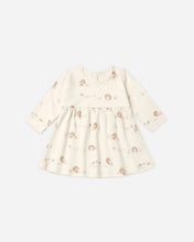 Load image into Gallery viewer, A long-sleeve baby dress in cream with a delicate brown and gold cartoon character print, displayed flat against a light gray background.
