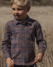 Load image into Gallery viewer, A young boy wearing a long-sleeve button-up plaid shirt in navy and red, standing in a dry grassy field.
