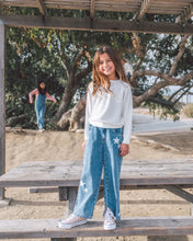 Load image into Gallery viewer, A young girl stands on a wooden picnic table outdoors under a pergola, wearing a white ribbed long-sleeve top and wide-leg blue jeans with white star patches on the knees, paired with white sneakers. Another child is visible in the background near trees.
