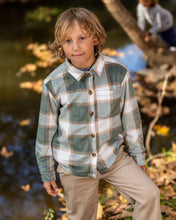 Load image into Gallery viewer, A young boy wearing a long-sleeve plaid button-up shirt in sage green, beige, and white, paired with khaki pants, standing outdoors near a stream with autumn leaves.
