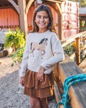 Load image into Gallery viewer, A young girl smiling while wearing a light gray knit sweater with a horse graphic and a brown tiered skirt, standing outdoors near a wooden railing.
