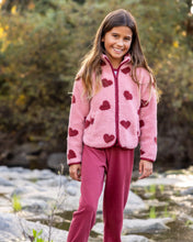 Load image into Gallery viewer, A young girl smiling outdoors wearing a pink fleece zip-up jacket with dark red heart patterns and matching pink pants.
