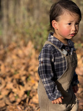 Load image into Gallery viewer, A young child standing outdoors on a ground covered with dry autumn leaves, wearing a blue and gray plaid flannel shirt under tan corduroy overalls.
