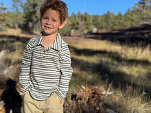Load image into Gallery viewer, A young boy with curly brown hair stands outdoors in a wooded setting, wearing a long-sleeve polo shirt with thin black and white horizontal stripes and tan pants.
