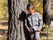 Load image into Gallery viewer, A young boy wearing a blue and gray plaid button-down shirt and gray pants leans against a large tree trunk in an outdoor park setting.
