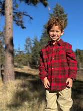 Load image into Gallery viewer, A young boy with curly light brown hair stands outdoors in a wooded area wearing a red and black plaid button-down shirt and khaki pants.
