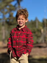 Load image into Gallery viewer, A young boy with curly light brown hair stands outdoors wearing a red and green plaid button-down shirt and beige pants, with a blurred forest and blue sky in the background.
