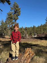 Load image into Gallery viewer, A young boy with curly red hair stands outdoors on a fallen log in a forest clearing, wearing a red and black plaid flannel shirt, tan pants, and dark sneakers under a clear blue sky.

