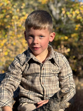 Load image into Gallery viewer, A young boy with short brown hair wearing a beige and brown plaid button-down shirt, sitting outdoors with a blurred background of yellow autumn leaves.
