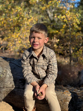 Load image into Gallery viewer, A young boy with short blond hair sits on a large log outdoors, wearing a beige and brown plaid button-up shirt and tan pants. He is holding small rocks in his hands, with a blurred background of trees and foliage in autumn colors.
