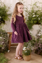 Load image into Gallery viewer, A young girl with long blonde hair stands in a styled indoor garden setting wearing a long-sleeve purple dress with a small gold floral pattern. She is wearing brown sandals and standing next to a small wicker basket filled with greenery.
