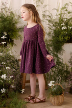 Load image into Gallery viewer, A young girl with long light brown hair stands in a garden-themed studio setting wearing a long-sleeve purple dress with a small gold floral pattern. She is wearing brown sandals and standing next to a wicker basket with greenery, with a wooden stool behind her against a cream-colored backdrop.
