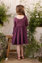 Load image into Gallery viewer, A young girl with long light brown hair is shown from behind, wearing a purple long-sleeve dress with a small floral pattern. She is standing in a styled indoor setting with a wooden stool and lush green potted plants against a cream-colored curtain background.
