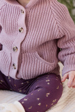 Load image into Gallery viewer, A close-up of a child wearing a dusty pink ribbed knit cardigan with brown buttons and dark purple patterned leggings, sitting on a cream blanket.

