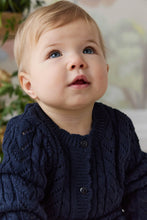 Load image into Gallery viewer, A close-up of a baby wearing a navy blue cable-knit cardigan sweater, looking upwards with a curious expression.

