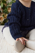 Load image into Gallery viewer, A close-up of a child wearing a navy blue cable-knit cardigan sweater, cream-colored leggings, and light beige boots, sitting on a textured cream blanket with a soft-focus floral background.
