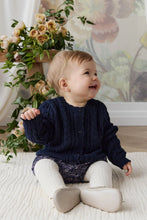 Load image into Gallery viewer, A baby sitting on a white textured rug wearing a navy blue cable-knit cardigan, patterned pants, and cream tights, with a floral arrangement and floral wallpaper in the background.
