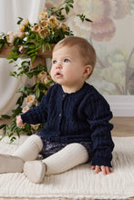 Load image into Gallery viewer, A baby sitting on a cream textured blanket, wearing a navy blue cable-knit cardigan, cream tights, and a sparkly navy skirt. The background features a floral arrangement and a soft floral wallpaper.
