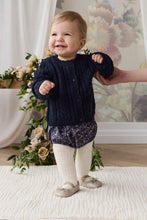 Load image into Gallery viewer, A smiling toddler stands on a white textured rug indoors, wearing a navy cable-knit cardigan, dark floral bloomers, cream tights, and beige shoes. A hand supports the child from the side, with flowers and a floral wall mural in the background.
