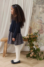 Load image into Gallery viewer, A young girl with long curly dark hair stands in profile wearing a navy cable-knit sweater, a dark floral print skirt, white tights, and navy flats. She is posed indoors next to a wooden bench and a floral arrangement against a backdrop of cream curtains and floral wallpaper.
