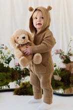 Load image into Gallery viewer, Child in a brown knit outfit with bear-ear hood holding Honey the Puppy plush among flowers and moss.
