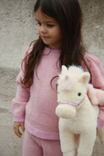 Load image into Gallery viewer, A young girl with long dark brown hair wearing a pink knit sweater and matching pants, holding a plush cream-colored stuffed horse against a textured gray wall.
