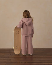 Load image into Gallery viewer, A child stands with their back to the camera, holding a wooden skateboard, wearing a matching dusty pink hoodie and wide-leg sweatpants against a textured beige wall and dark wood floor.
