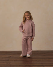 Load image into Gallery viewer, A young girl stands on a dark wood floor against a beige textured wall, wearing a matching dusty rose pink two-piece loungewear set consisting of a pullover hoodie and wide-leg pants.
