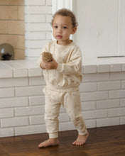 Load image into Gallery viewer, A toddler stands barefoot indoors in front of a white brick fireplace, wearing a cream-colored long-sleeve sweatshirt and matching sweatpants with a subtle allover animal print. The child is holding a small pinecone.
