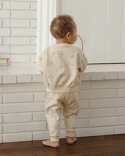 Load image into Gallery viewer, A baby standing with their back to the camera, wearing a matching beige two-piece pajama set with a subtle teddy bear print, in front of a white brick fireplace.
