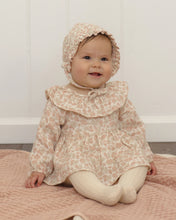 Load image into Gallery viewer, A baby wearing a light beige and pink floral print long-sleeve dress with a large ruffled collar, matching bonnet, and cream tights, sitting on a pink quilted blanket against a white paneled wall.
