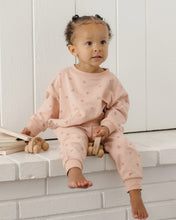Load image into Gallery viewer, A toddler girl sits barefoot on a white brick ledge wearing a light pink long-sleeve sweatshirt and matching jogger pants with a small heart pattern. She is holding a wooden toy, with a stack of wooden toys on the left, against a white paneled wall background.
