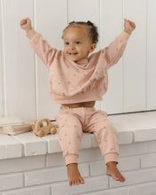 Load image into Gallery viewer, A toddler girl wearing a matching dusty pink two-piece loungewear set with a subtle heart pattern, sitting on a white brick ledge with her arms raised in excitement.
