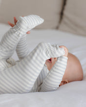 Load image into Gallery viewer, A baby lying on a white bed, wearing a long-sleeve footed sleeper with thin gray and white stripes.
