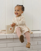 Load image into Gallery viewer, A smiling toddler girl sits on a white brick ledge holding a small beige book. She is wearing a cream long-sleeve dress with a delicate floral pattern, pink tights, and tan Mary Jane shoes against a white paneled wall background.
