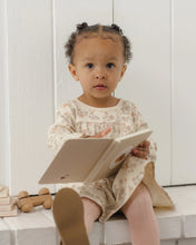 Load image into Gallery viewer, A toddler girl with braided hair sits on a white bench reading a book, wearing a cream floral long-sleeve dress and pink tights.
