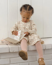 Load image into Gallery viewer, A toddler girl sits on a white brick bench reading a book, wearing a cream floral long-sleeve dress, pink ribbed tights, and brown Mary Jane shoes.
