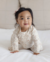 Load image into Gallery viewer, A baby girl wearing a cream-colored floral long-sleeve footless sleeper sits on a white bed, looking towards the camera.
