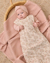 Load image into Gallery viewer, A baby lying on a pink textured blanket inside a woven basket, wearing a pale pink knotted beanie and a cream floral long-sleeve swaddle blanket.
