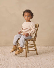 Load image into Gallery viewer, A toddler sits on a rustic wooden chair wearing a cream and beige striped knit sweater, light gray pants, and tan shoes against a neutral beige backdrop.
