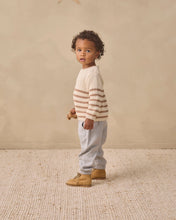 Load image into Gallery viewer, A toddler standing in profile wearing a cream knit sweater with brown horizontal stripes, light gray pants, and tan boots, set against a warm beige studio backdrop.
