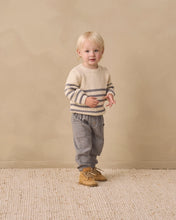 Load image into Gallery viewer, A toddler standing against a warm beige studio backdrop, wearing a cream knit sweater with dark horizontal stripes, gray pants, and tan lace-up boots.
