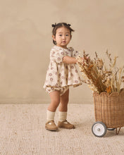 Load image into Gallery viewer, A toddler girl stands in a studio setting wearing a cream-colored short-sleeve romper with a brown floral pattern, paired with cream socks and brown shoes. She is positioned next to a wicker basket filled with dried flowers on a textured beige rug.
