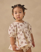 Load image into Gallery viewer, A toddler girl with two buns in her hair wears a cream-colored cotton dress with a brown floral pattern, standing against a solid beige background.
